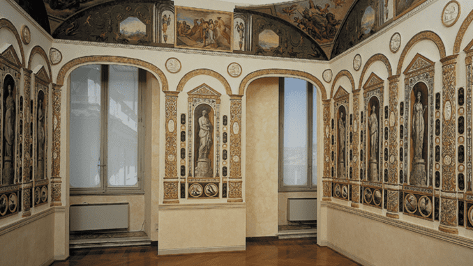 Ornate historic room with high arched windows, frescoes, bas-relief sculptures, and polished wood floor.