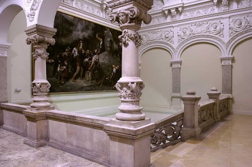 Ornate indoor space with arches, columns, detailed carvings, water basin, and large painting on wall.