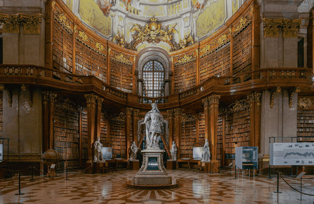Ornate library with central statue, marble floor, and high bookshelves illuminated by large arched window.