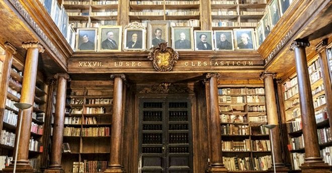 Ornate library interior with tall wooden shelves, large columns, an upper gallery, and portraits.
