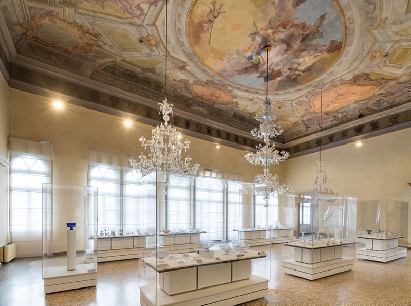 Ornate museum interior with decorated ceiling fresco, chandeliers, display cases, and large windows.