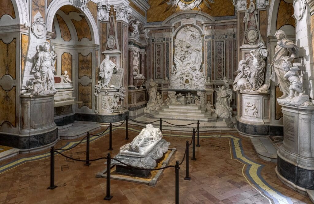 Ornately decorated room with intricate marble sculptures, central reclining figure veiled, black rope barrier.