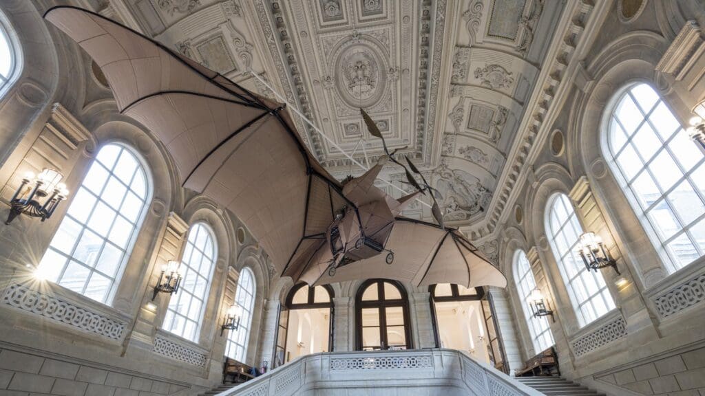 ornithopter suspended in ornately detailed hall with arched windows and grand staircase