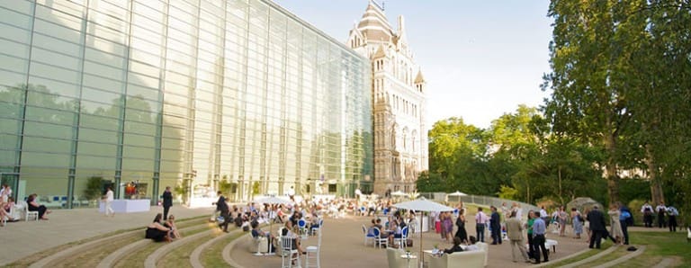Outdoor gathering in front of modern glass building and historic structure with people and white chairs.