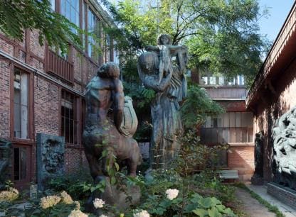 Outdoor sculpture garden with large statues between brick buildings surrounded by greenery.