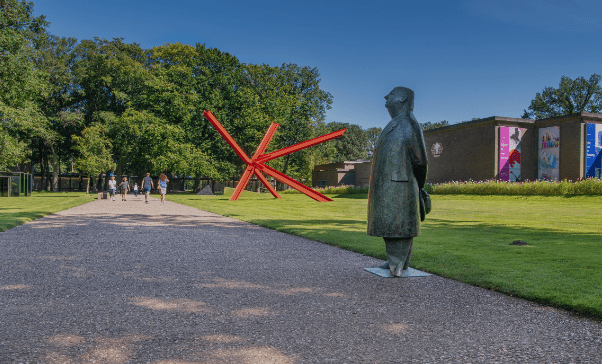 Outdoor area with a red sculpture, a statue, people walking on a paved path, and colorful banners.