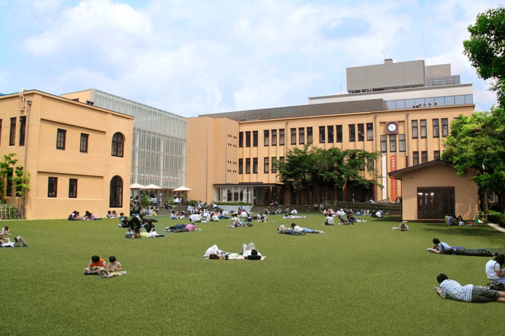 People relaxing on a large grassy field in front of modern beige buildings under clear skies.