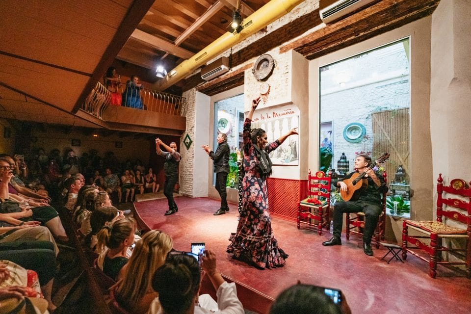 Performers dance on stage in rustic indoor venue with musician and attentive audience.