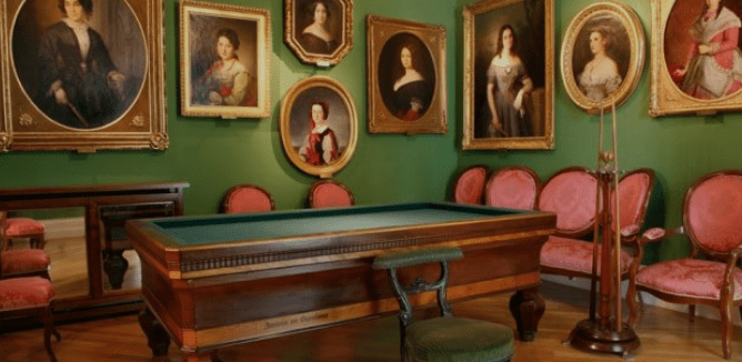 Room with period portraits, green walls, pink-upholstered vintage chairs, ornate table, and coat rack