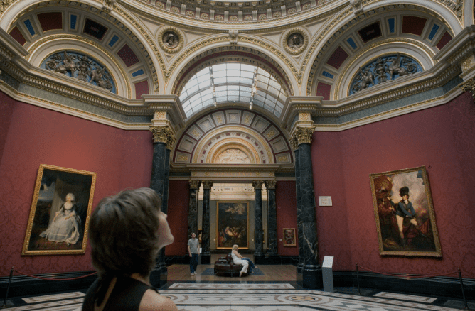 Person admiring ornate ceiling and classical architecture in an art gallery with framed paintings.
