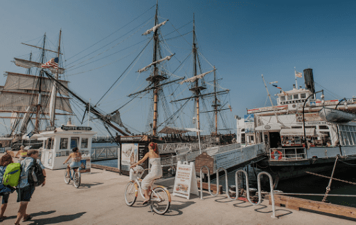 Pier with historical ships and modern ferry, people walking and cycling, ticket booth nearby.
