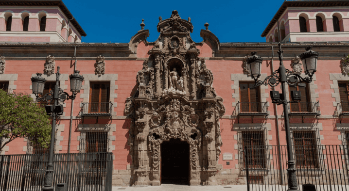 Ornate baroque-style entrance of a pink historic building with sculptures, balconies, lamps, and a tree.