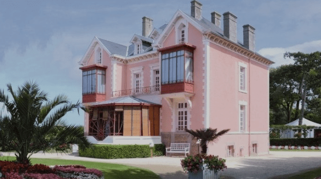 Two-story pink house with mansard roof, large windows, conservatory, garden, bushes, flowers, palm trees.