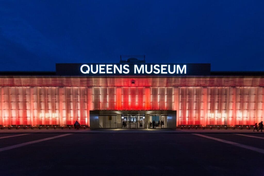 Illuminated facade of Queens Museum at night with warm lights and people near the entrance.