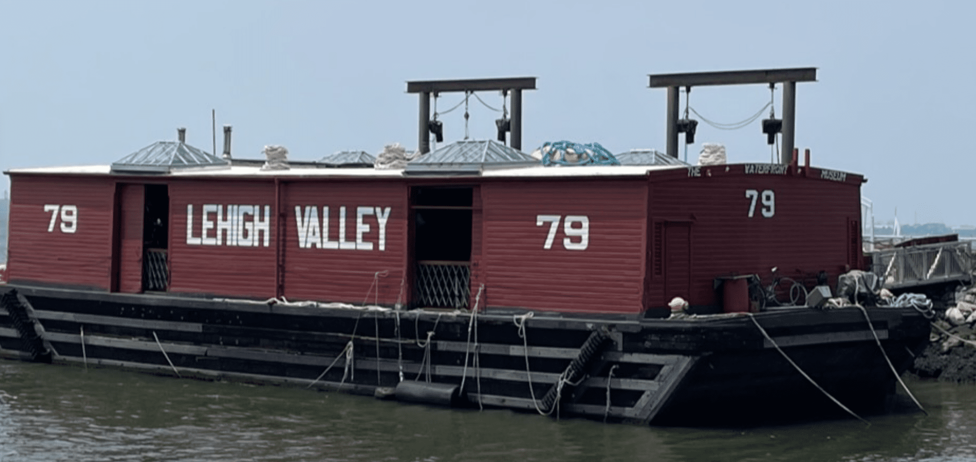 Red barge with "LEHIGH VALLEY" and "79" docked in water with equipment on its roof.
