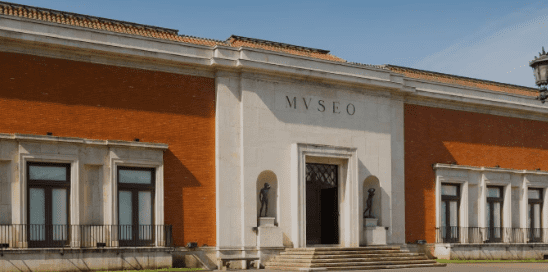 Large classical building with red-brick walls, stone entrance labeled "MVSEO," statues, and steps.