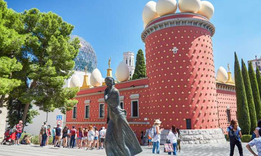 Tourists gather outside a unique red building with egg sculptures and a prominent statue in front.