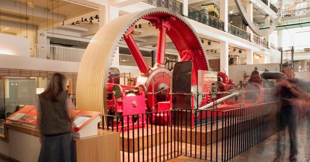 Red industrial machine exhibit with a large gear in a modern museum, viewed by visitors.