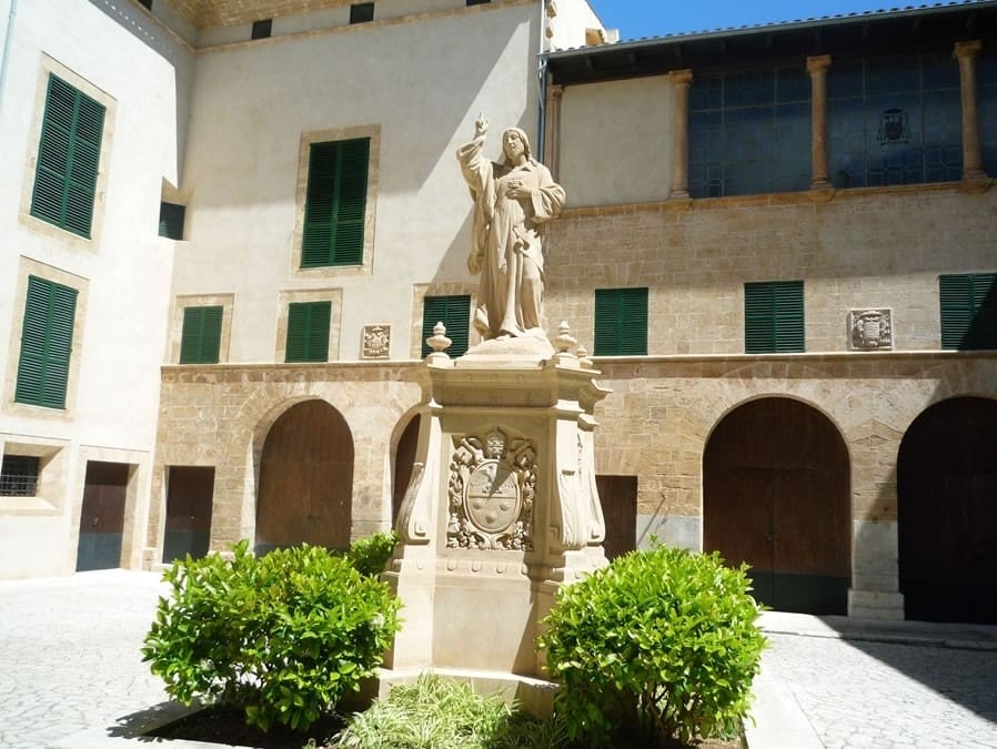 Statue of a robed figure on a pedestal in a courtyard with arched doorways and green shuttered windows.