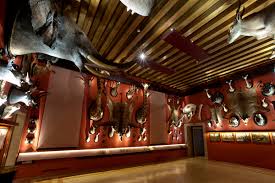 Room with wooden-beamed ceiling, red walls, mounted animal heads, and display cases with framed pictures