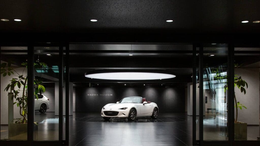 Showroom with a white convertible car and "Mazda Museum" sign on a minimalist black wall.