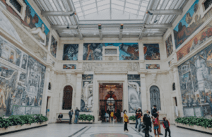 Spacious art museum interior with high glass ceiling, murals, ornamental door, and visitors.