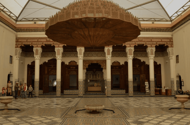 Spacious courtyard with Islamic architecture, ornate ceiling, mosaic floor, columns, and visitors.