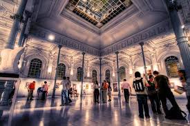 Spacious hall with high ceilings, arched windows, columns, and people observing an exhibition.