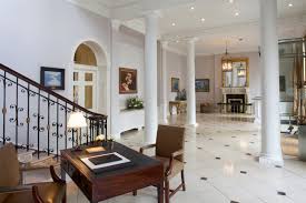 Spacious interior with marble floors, white columns, a wooden desk, staircase, and chandelier.