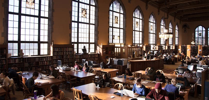 A spacious library with large arched windows, wooden tables, and patrons reading and studying.