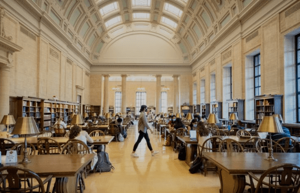 Spacious library with high ceiling, arched windows, wooden tables, lamps, people studying or working.