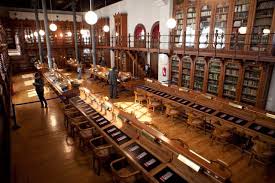 A spacious library with wooden furniture, long tables, bookshelves, and large round light fixtures