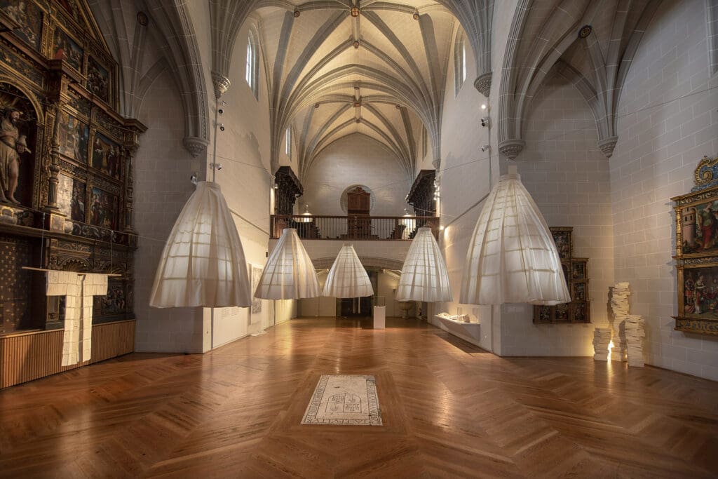 Spacious room with ornate ceiling, large cone-shaped light fixtures, wooden floors, and historical artworks.