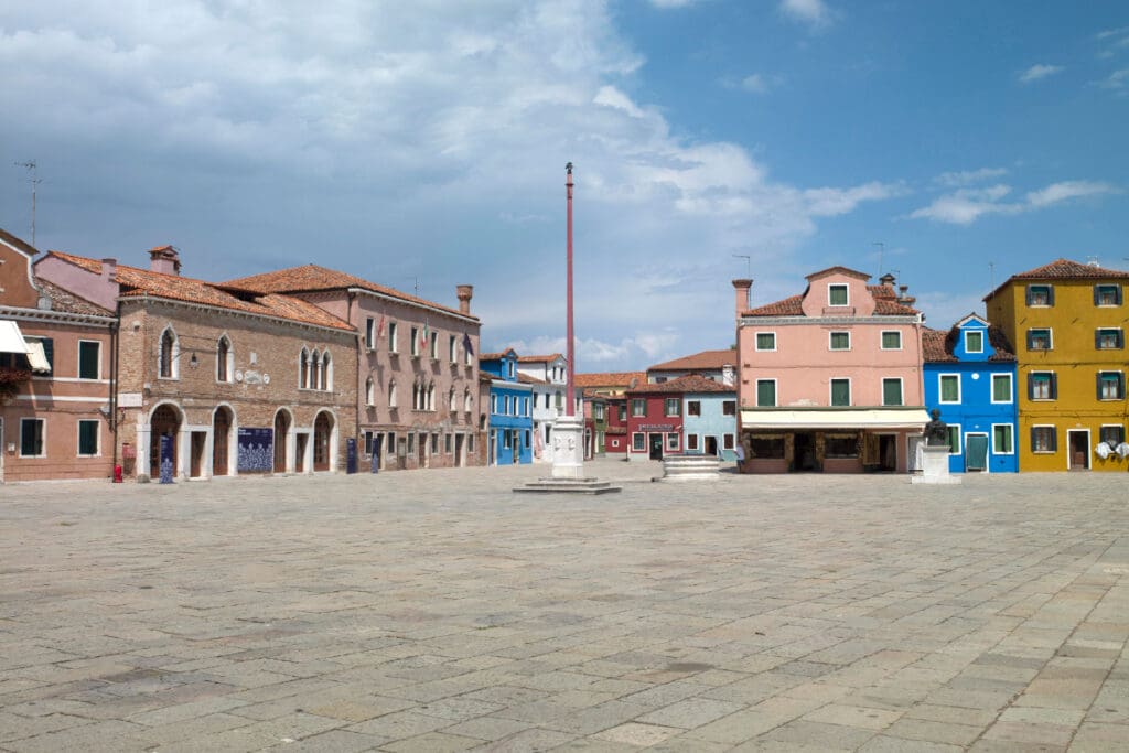 Spacious, paved town square with colorful buildings, tall red pole, and partly cloudy sky.