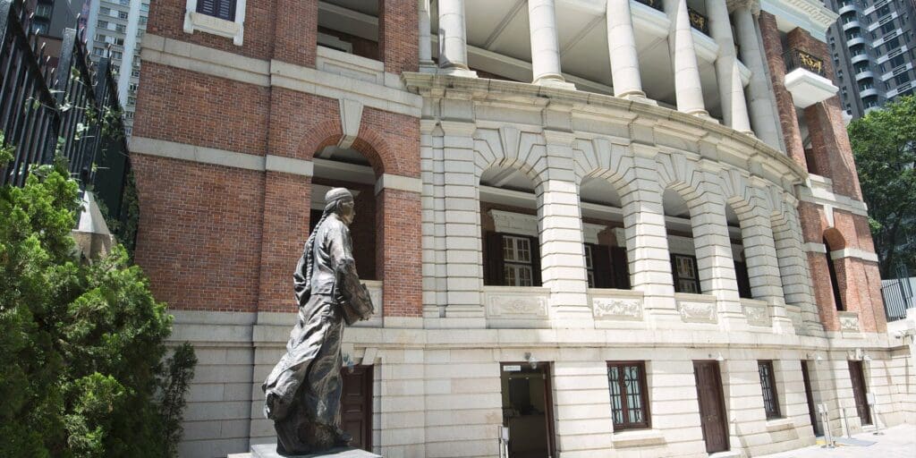 Statue in front of historic red brick building with arched openings and surrounding urban landscape.
