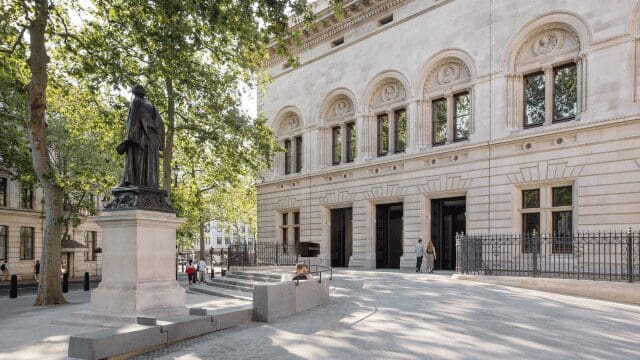 Statue on a pedestal in a tree-lined courtyard with an ornate building and people in the background