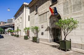 Stone building with archway entrance, flag above, potted trees, paved path, urban background.