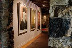 Corridor with stone walls, wooden ceiling, framed portraits on left, leading to dimly lit area.