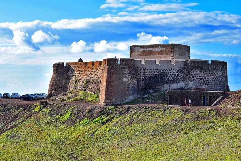 Stone fort on grassy hill with cars and people, under a partly cloudy sky background.