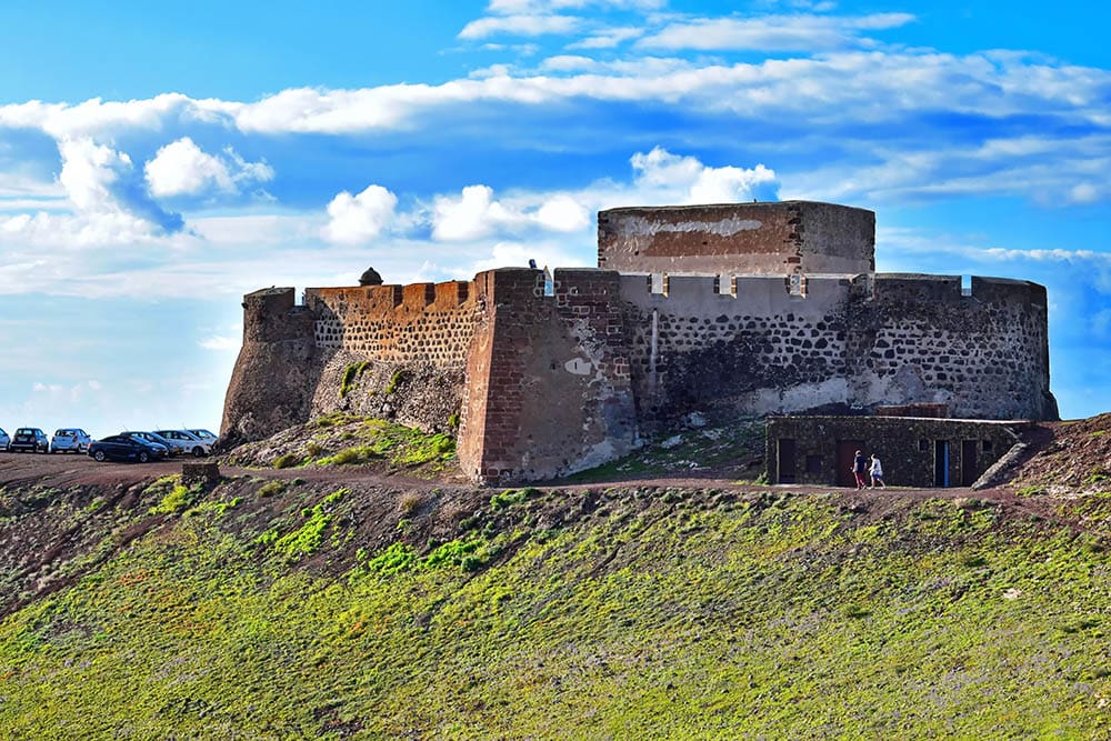 Stone fort on grassy hill with cars and people, under a partly cloudy sky background.
