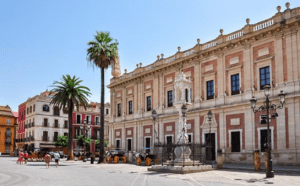 Sunny city square with a historic neoclassical building, palm trees, statue, and horse-drawn carriages.