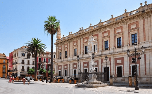 Sunny city square with a historic neoclassical building, palm trees, statue, and horse-drawn carriages.