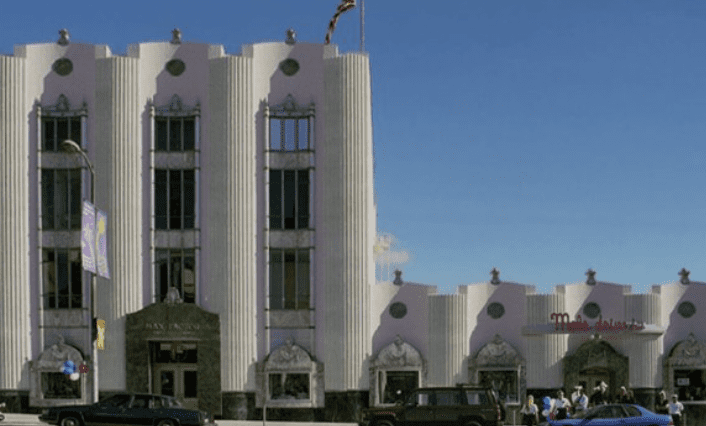 Three-story Art Deco building with vertical fluted facade, gallery sign, flagpole, cars, and people.