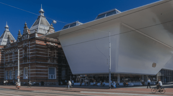 Traditional brick building with turrets beside a modern white structure, with pedestrians and cyclists.