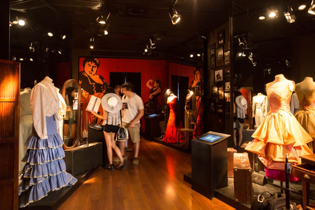 Exhibit of traditional Spanish costumes with visitors examining displays in a dimly lit museum.