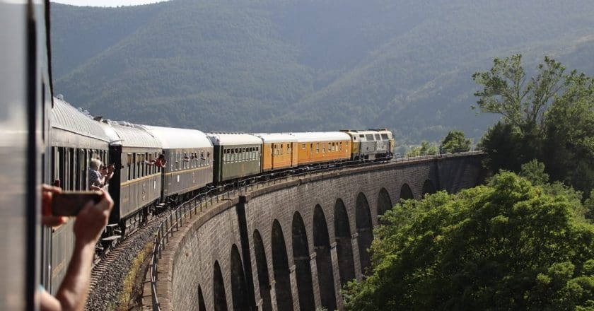 Train with passengers capturing scenic views crosses an arched viaduct amidst lush green landscape.