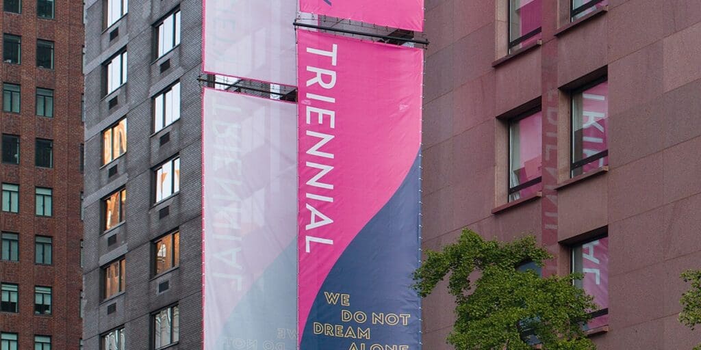 Vertical banners on a modern building displaying "TRIENNIAL" and "WE DO NOT DREAM ALONE."