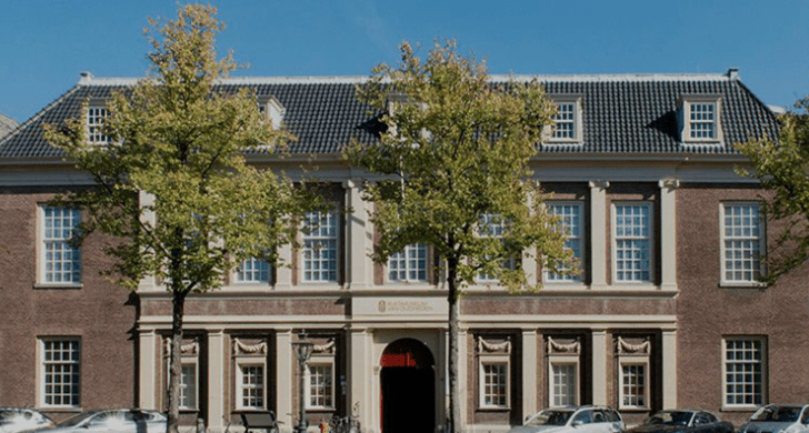 A large, two-story brick building with classical architecture, tall windows, trees, and parked cars.
