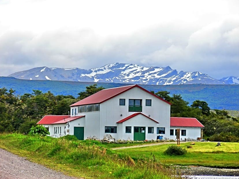 Large two-story building with red roof in natural landscape, surrounded by greenery, snowy mountains.