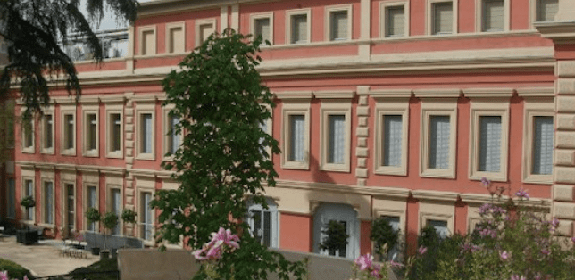 Large two-story red and beige building with windows, a tree, and surrounding flowering plants.